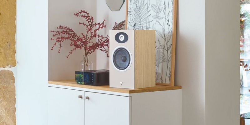 Diffusore Focal Theva N°1 in legno chiaro appoggiato su una credenza bianca accanto a un vaso di fiori e a un quadro